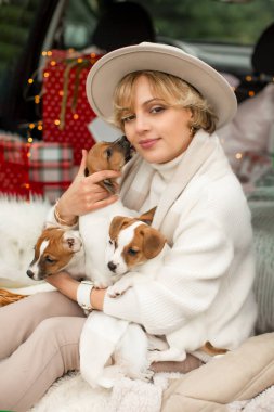Hafif giysili ve şapkalı bir kız, Jack Russell Terrier köpeğiyle bir arabanın bagajında oturuyor. Noel tatiller için bir Noel ağacı alıyor..
