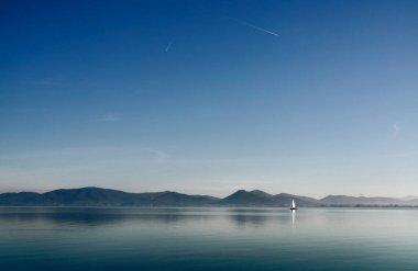 One sail boat in the calm waters of Lake Trasimeno , Italy ,in the background small towns clipart