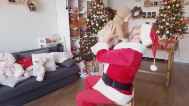 Kırmızı ve beyaz kıyafetli Noel Baba, bebekle süslenmiş Noel ağaçlarının arka planında parlayan çelenkler ve hediye kutularıyla oynar.