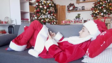 Kırmızı-beyaz takım elbiseli genç Noel Baba, yeni yıl süslemelerinin arka planında koltukta yatıyor ve telefon için tebrik ediyor.
