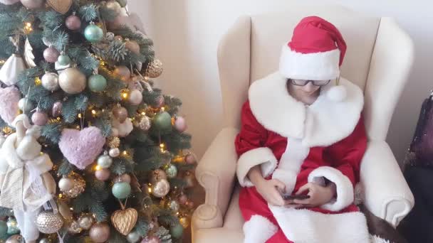 Молодой Санта Клаус Красно Белом Костюме Сидит Кресле Фоне Рождественской — стоковое видео