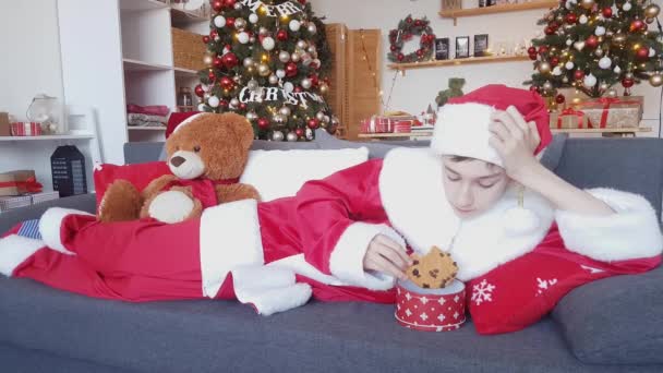 若いサンタクロースは 装飾されたクリスマスツリーとテディベアの背景にソファの上に横たわるレーズンとクリスマスジンジャーブレッドを食べます — ストック動画