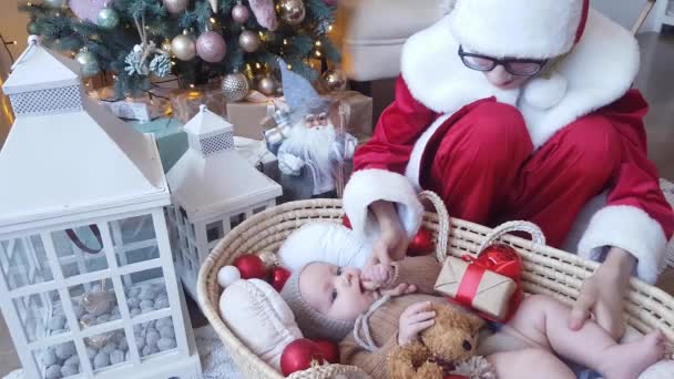 年轻的圣诞老人身穿漂亮的红白相间的衣服 戴着眼镜 送给躺在柳条篮里的婴儿一份礼物 礼物的背景是新年的装饰品 — 图库视频影像