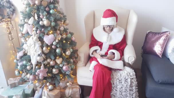 身穿红白相间衣服的圣诞老人坐在圣诞树前的扶手椅上 用电话向圣诞老人道贺 — 图库视频影像