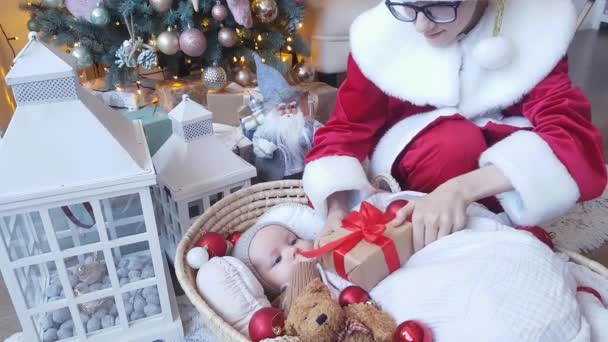 美しい赤と白の衣装を着た若いサンタクロースは 眼鏡をかけて 新年の装飾の背景にある邪悪なバスケットに横たわっている赤ちゃんに贈り物を与えます — ストック動画