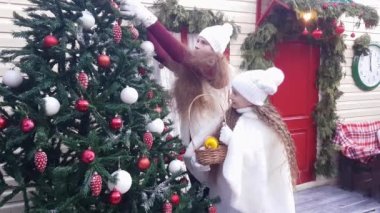 Şirin çocuklar, sarı kıvırcık saçlı, beyaz şapkalı ve kürklü kızlar bahçedeki Noel ağacını renkli toplarla süslüyor.