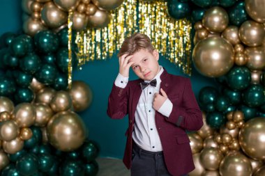 Siyah pantolon giymiş bir çocuk papyonlu beyaz gömlek ve bordo ceketli yeşil arka planda altın kemer ve yeşil balonlar içinde duruyor.