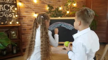 Sarışın okul çocukları - bir oğlan ve bir kız, tahtaya rengarenk pastel kalemler çiziyor. Taze çiçeklerin arka planına, tuğla bir duvara ve yazıya karşı.!