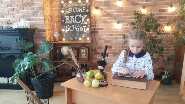 一个女孩正在学习用粉笔写在写字板上 靠着显微镜 放大镜 苹果和梨子坐在写字台前 在题词后面 回学校去 — 图库视频影像