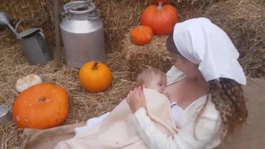 A young mother is breastfeeding her baby, sitting on a linen cloth in stacks of straw against the backdrop of pumpkins and rural accessories