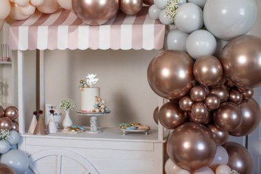 Kapalı bir dolabın arka planında, kahverengi, bej ve gri helyum balonları ve çiçeklerle süslenmiş sokak satıcıları. Sepetin üzerinde bir pasta var. Üstünde de 