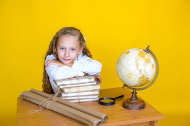 A blond schoolgirl sits at a desk  on a yellow background. There is a globe, books, scrolls and magnifying glass