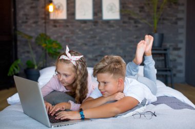 Fair-haired children - a boy and a girl study and play at a laptop, lying on the bed