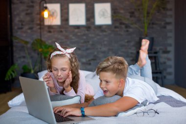 Fair-haired children - a boy and a girl study and play at a laptop, lying on the bed