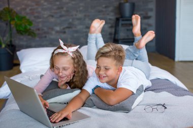 Fair-haired children - a boy and a girl study and play at a laptop, lying on the bed