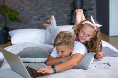 Fair-haired children - a boy and a girl study and play at a laptop, lying on the bed