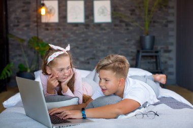 Fair-haired children - a boy and a girl study and play at a laptop, lying on the bed