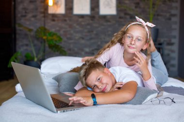 Fair-haired children - a boy and a girl study and play at a laptop, lying on the bed
