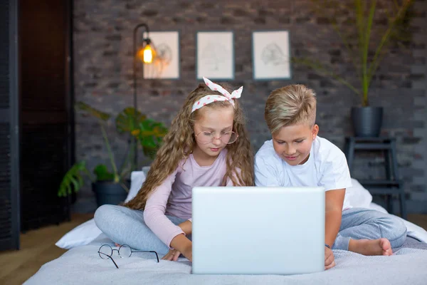 Fair-haired children - a boy and a girl study and play at a laptop, sitting on the bed