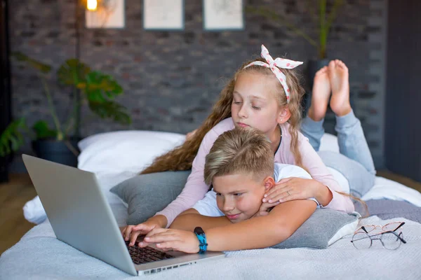 Fair-haired children - a boy and a girl study and play at a laptop, lying on the bed