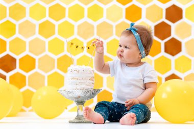 Dişi bir bebek bir pastanın yanına oturur ve elleriyle bir parça yer. Arka planda sarı bal peteği ve balon tasarımı var. Kek konsepti.