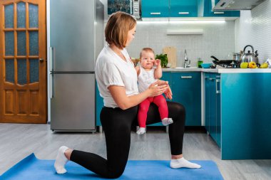 Yoga evde. Kafkas kökenli genç bir anne spor yapıyor ve elinde bir bebek tutuyor. Evde çocuklarla fitness konsepti