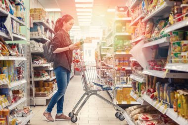 Genç ve güzel bir kadın market arabasını yuvarlıyor ve süpermarketten ürünler seçiyor. Işık.