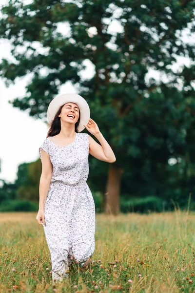 夏天的时间 一个穿着连衣裙 头戴草帽的快乐的年轻女子站在田边 后面是一棵大树 垂直方向 — 图库照片