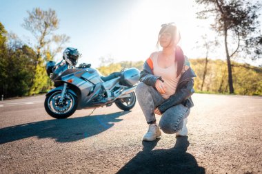 Dünya Motosiklet Günü. Kolunda dövmeleri olan, deri ceket giyen ve poz veren genç bir kadın. Arka planda yol ve motosiklet var. Motosiklet yerel seyahat kavramı.