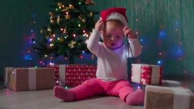 Komik küçük bir kız Noel ağacının yanında oturur ve Noel şapkasını çıkarır. Ağır çekim. Kış tatili kavramı.