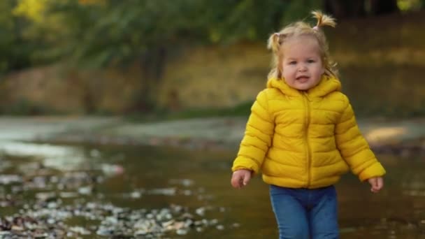 黄色のゴム製のブーツとジャケットの小さな面白い女の子が川岸を歩いています 傾いて スローモーション — ストック動画