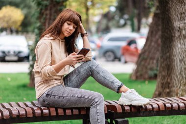 İletişim konsepti. Güzel bir kadın parkta bir bankta oturuyor ve akıllı bir telefonda daktilo yazıyor. Açık havada. Ücretsiz kablosuz ağ alanı.