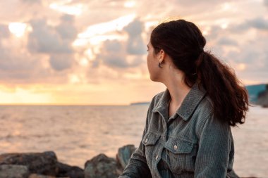 Genç bir kadın deniz kenarında tek başına oturur ve gün batımını seyreder. Arkadan bak. Psikoloji ve akıl sağlığı kavramı.