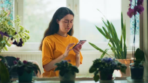 白人の若い女性は スマートフォンの窓の近くに自宅で座ってチャット 自然植物とのインテリア リアルタイムだ ソーシャルメディア中毒と携帯アプリの概念 — ストック動画