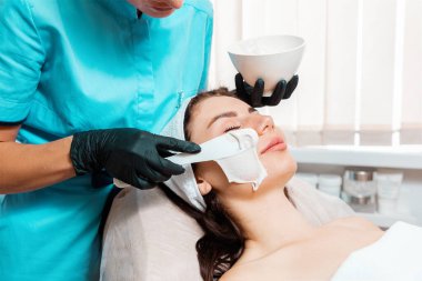 Salon prosedürü. Lateks eldivenli güzellik uzmanı güzel kadının yüzüne spatula gençleştirme maskesi takıyor. Kapatın. Profesyonel kozmetoloji kavramı.