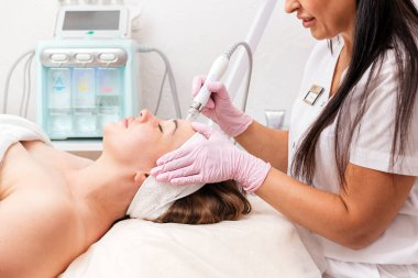 Güzellik salonu. Plastik eldivenli bir kozmetik uzmanı yüzüne su soyma prosedürü uygular. Arka planda bir kozmetoloji cihazı var. Profesyonel kozmetoloji ve güzellik kavramı.