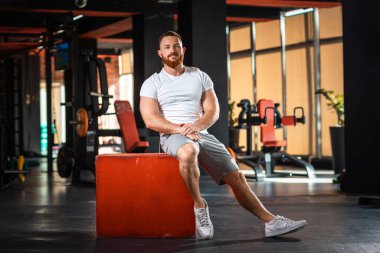 Yakışıklı atletik sakallı adam modern spor salonunda bir küpün üzerinde oturuyor. Zindelik ve refah kavramı.