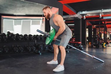 Sakallı bir sporcu spor salonunda halter kaldırarak antrenman yapıyor. Çapraz eğitime ayak uyduruyorum. Spor anlayışı.