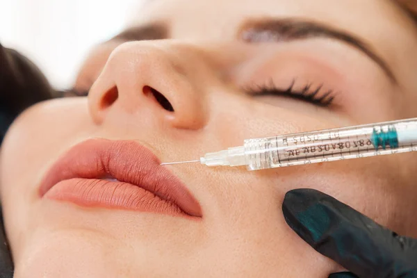 Aesthetic medicine and cosmetology. Cosmetologist in black latex gloves makes with syringe injects of a medical in lips. Close up of face. Concept of plastic surgery.