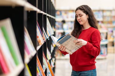 Kafkasyalı genç bir kadın elinde kitaplarla rafların arkasında duran bir kitap okuyor. Eğitim ve kitap satın alma kavramı.