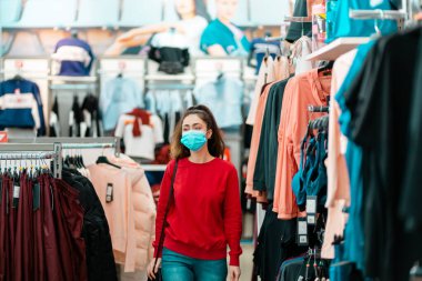 Halka açık yerlerde antiviral koruma. Tıbbi maskeli genç ve güzel bir kadın mağazadan kıyafet seçiyor. Virüs salgını sırasında alışveriş kavramı.