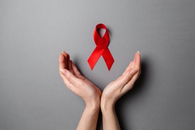 Kadının elleri, AIDS 'le mücadelenin sembolü olan kırmızı kurdeleyi çerçeveletiyor. Gri arka plan. Düz yatıyordu. Uzayı kopyala Dünya AIDS Günü konsepti.