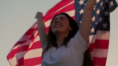 Şarkı söyleyen, dans eden ve Amerikan bayrağını sallayan mutlu genç bir kadın. Günbatımı gökyüzünün arka planında. Alt manzara. Ağır çekim. Bağımsızlık Günü kavramı