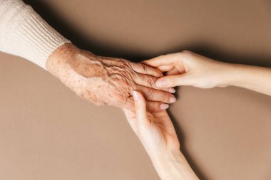 Genç bir kadının elleri yaşlı bir büyükannenin buruşuk ellerini tutar. Bej arka plan. Düz yatıyordu. Yaşlılarla ilgilenme kavramı.