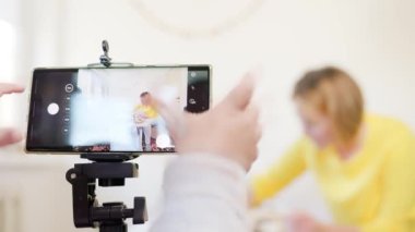 Video yayını. Küçük bir çocuk video kaydedilirken parmaklarını akıllı telefon ekranına bastırıyor. Arka planda kadın masada kurabiye pişiriyor..