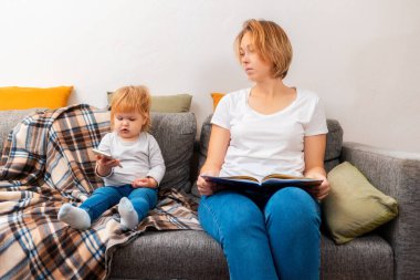 Anne elinde bir kitapla kanepede oturuyor, ve küçük kızı telefonda oyun oynuyor. Çocukların online oyunlara olan bağımlılığı..