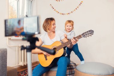 Mutlu genç anne ve küçük çocuğu cep telefonunda canlı yayında gitar çalıyorlar. Video yayını ve çevrimiçi öğrenme kavramı.