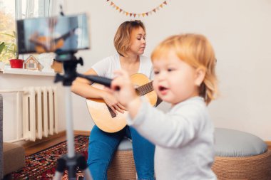 Genç bir anne gitar çalıyor ve akıllı telefondan canlı yayın yapıyor. Ön planda bulanık bir çocuk. Video akışı ve çevrimiçi öğrenme kavramı.