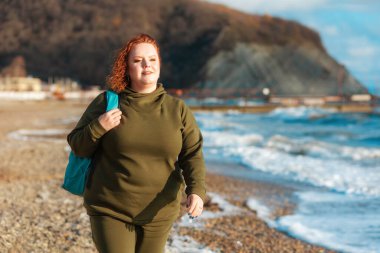 Sırt çantalı, kumsalda yürüyen iri yarı, güzel bir kadın. Arka planda bir okyanus ve bir dağ var. Seyahat kavramı.