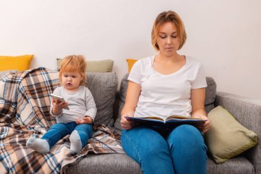 Bir kadın kanepede oturuyor ve kitap okuyor. Küçük bir çocuk onun yanında oturuyor ve telefonda oynuyor. Aile rahatla.